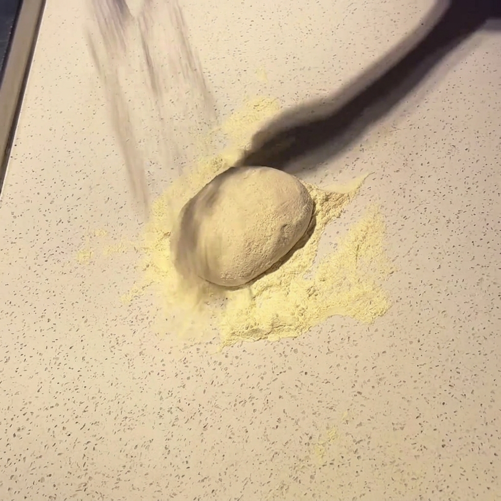 Eine Kugel Pizzateig wird auf einer hellen Arbeitsfläche mit Mehl bestäubt.