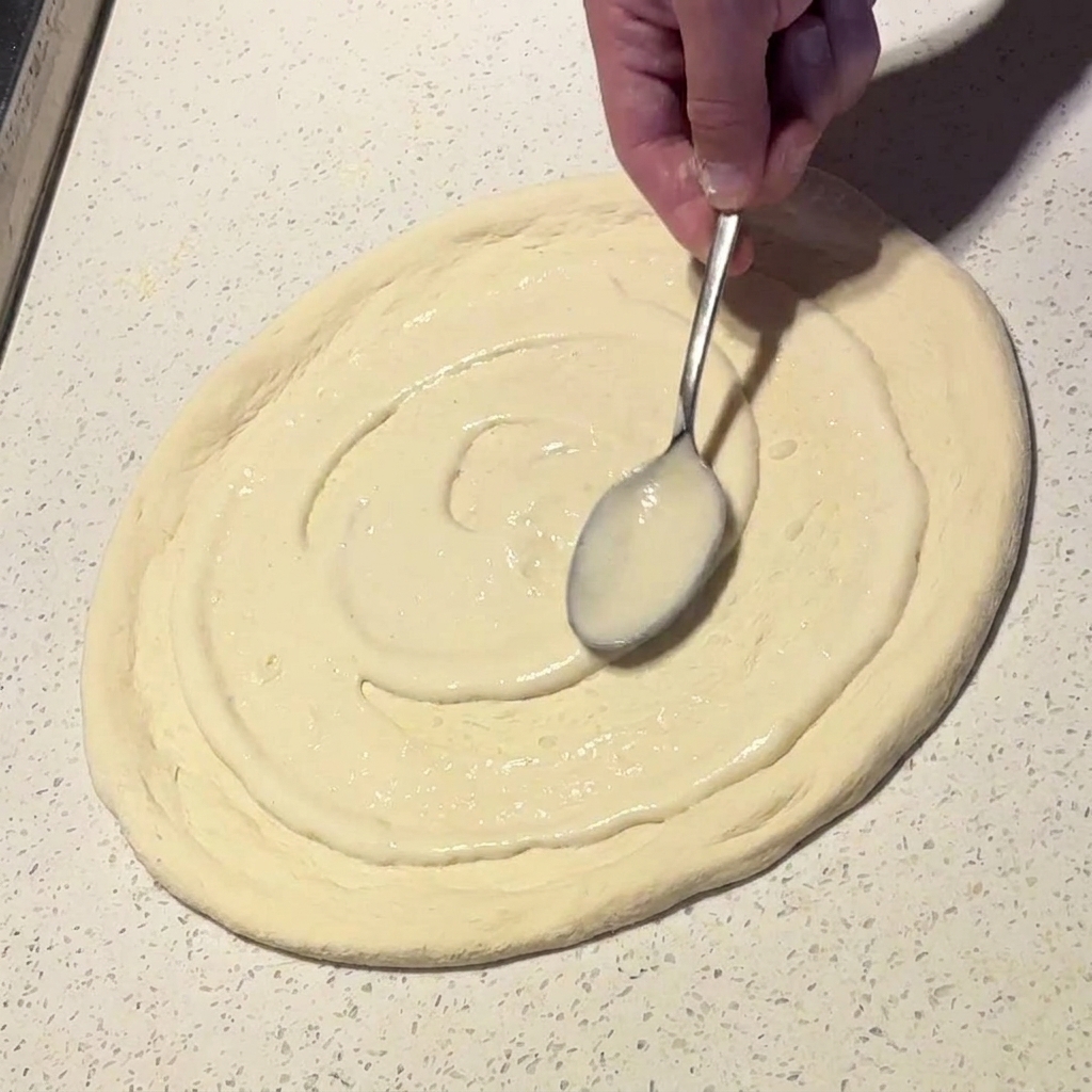 Ein Metalllöffel verteilt cremige weiße Sauce in kreisenden Bewegungen auf einem Pizzateigboden.
