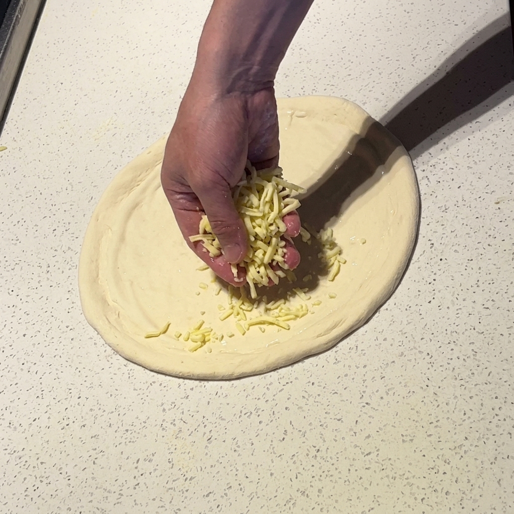 Eine Hand streut geriebenen Mozzarella über den mit weißer Sauce bestrichenen Pizzateig.