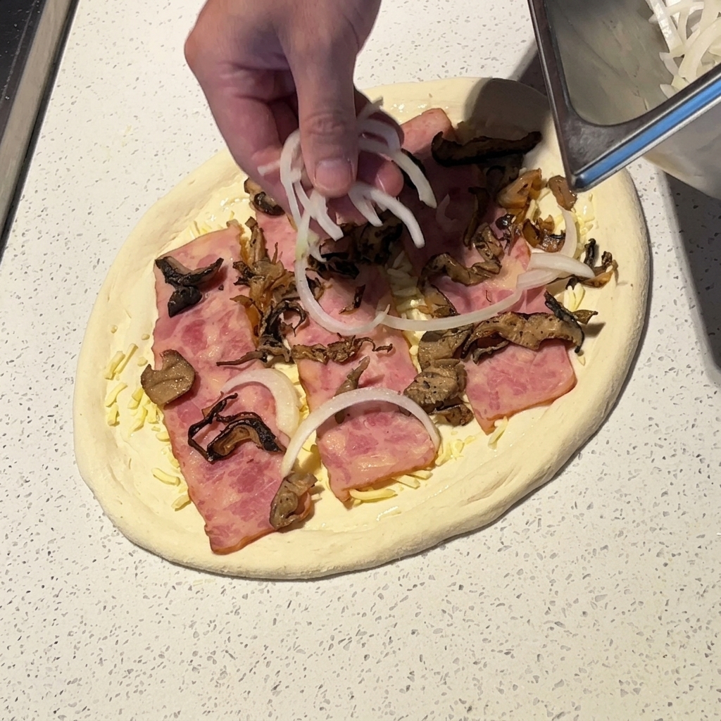 Eine Hand verteilt dünne weiße Zwiebelringe auf der Pizza.