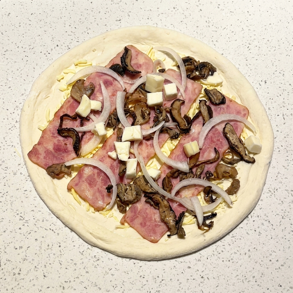Die fertig belegte rohe Pizza mit Speck, Pilzen, Zwiebeln und frischen Mozzarella-Würfeln.