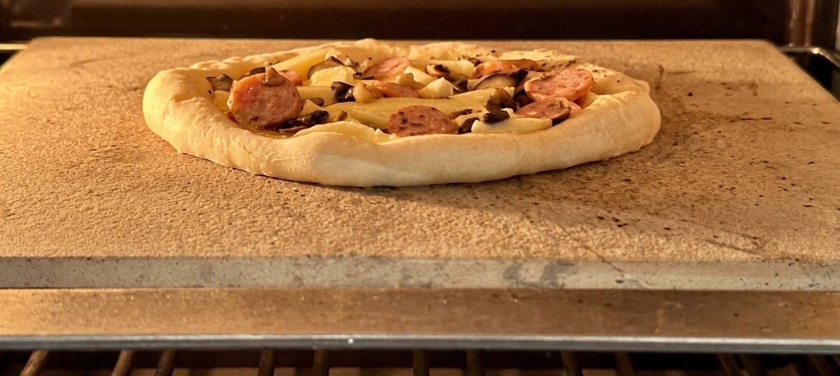 Eine fertig belegte Pizza mit Wurst und Pilzen auf einem Backstein im heißen Ofen.