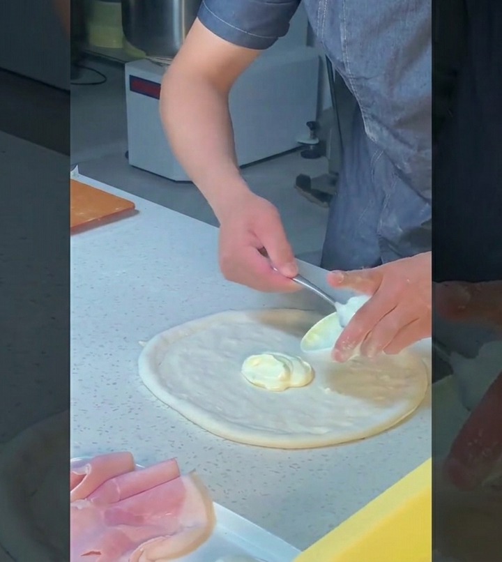 Ein Koch verteilt mit der Rückseite eines Löffels weiße Sauce kreisförmig auf dem Pizzateig.