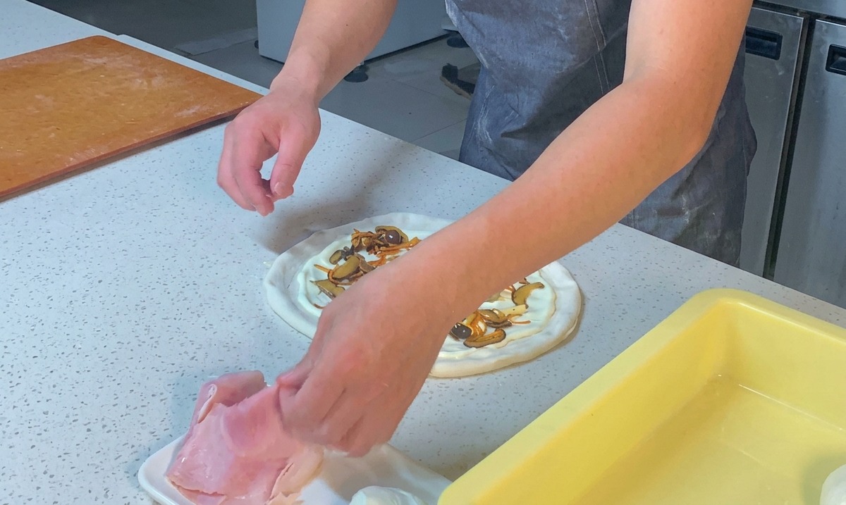Hände zupfen Schinkenstücke und verteilen sie auf der Pizza.