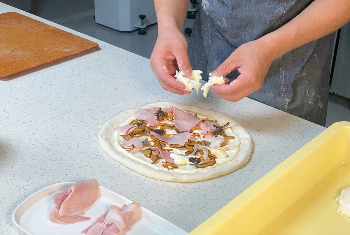 Hände zupfen frischen Mozzarella über die Pizza mit Schinken und Pilzen.