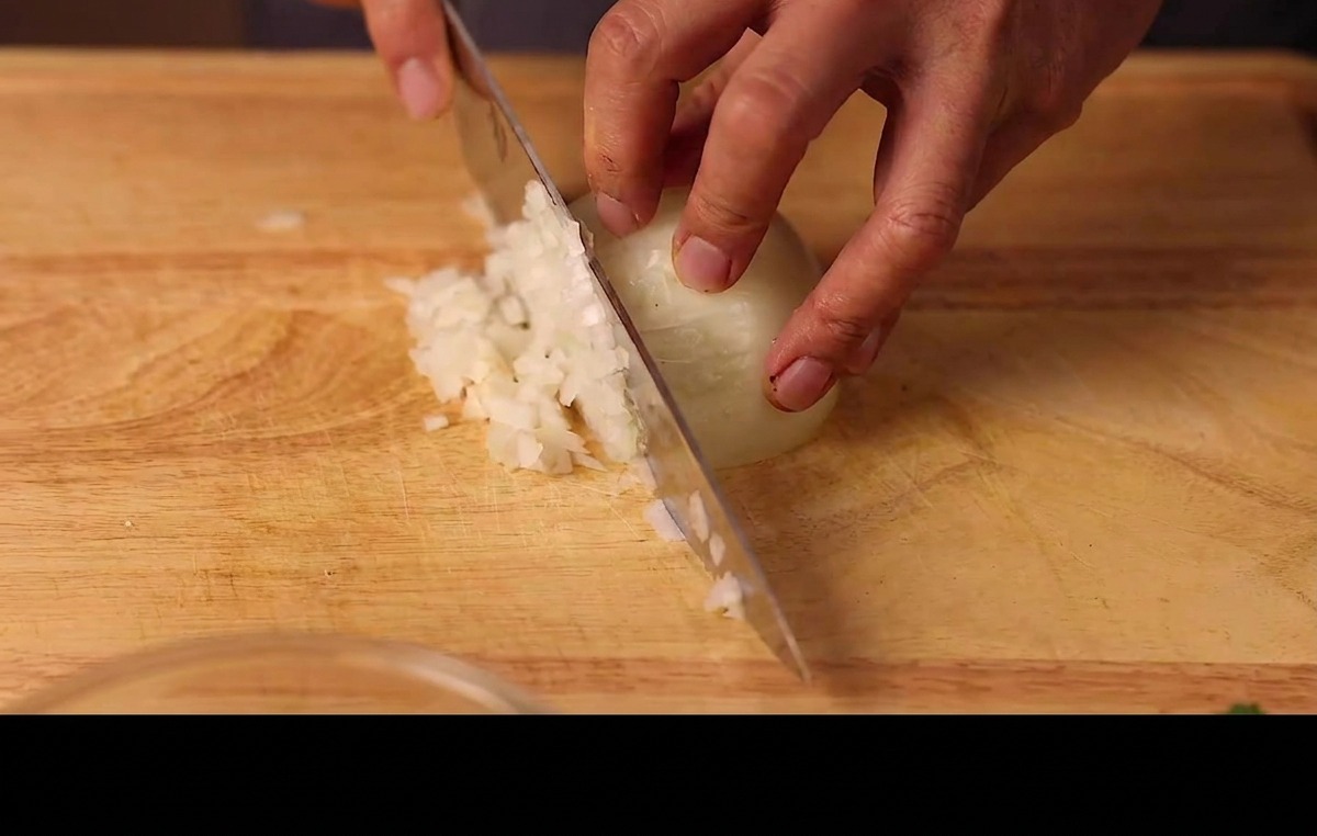 Hände würfeln eine halbe weiße Zwiebel in feine Stücke auf einem Schneidebrett aus Holz.
