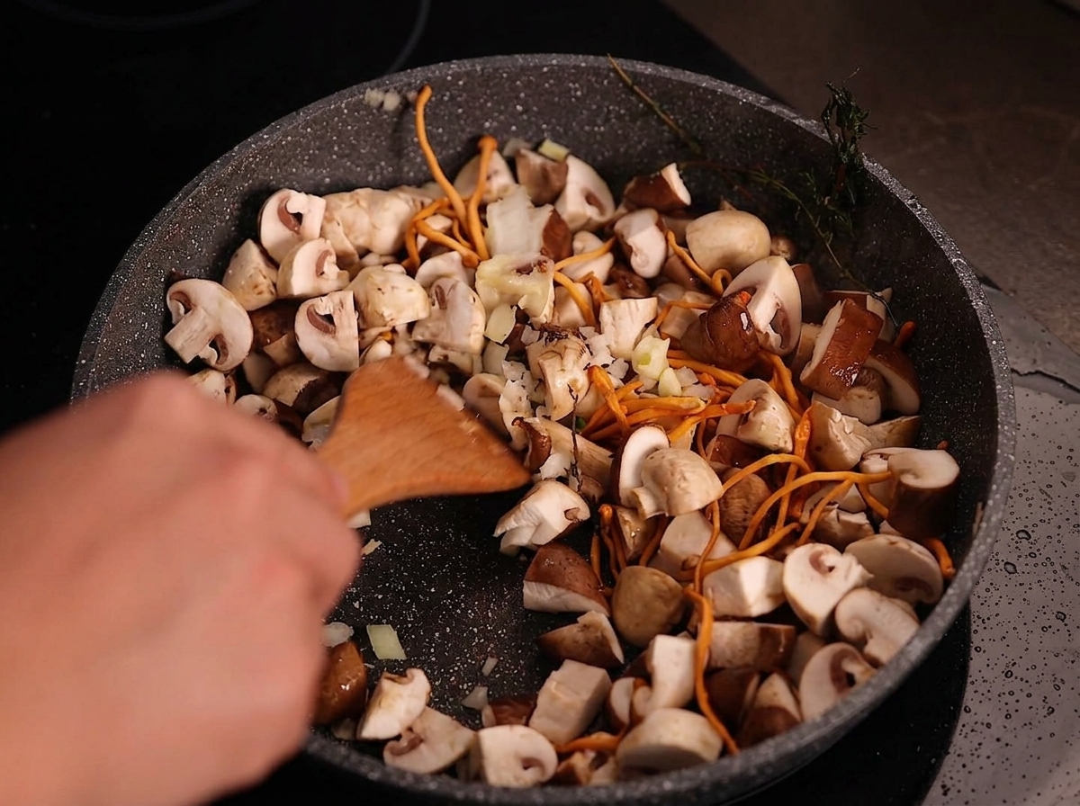 Ein Holzspatel rührt gehackte weiße Zwiebeln und geschnittenen Knoblauch in eine Pfanne mit gebratenen Pilzen.