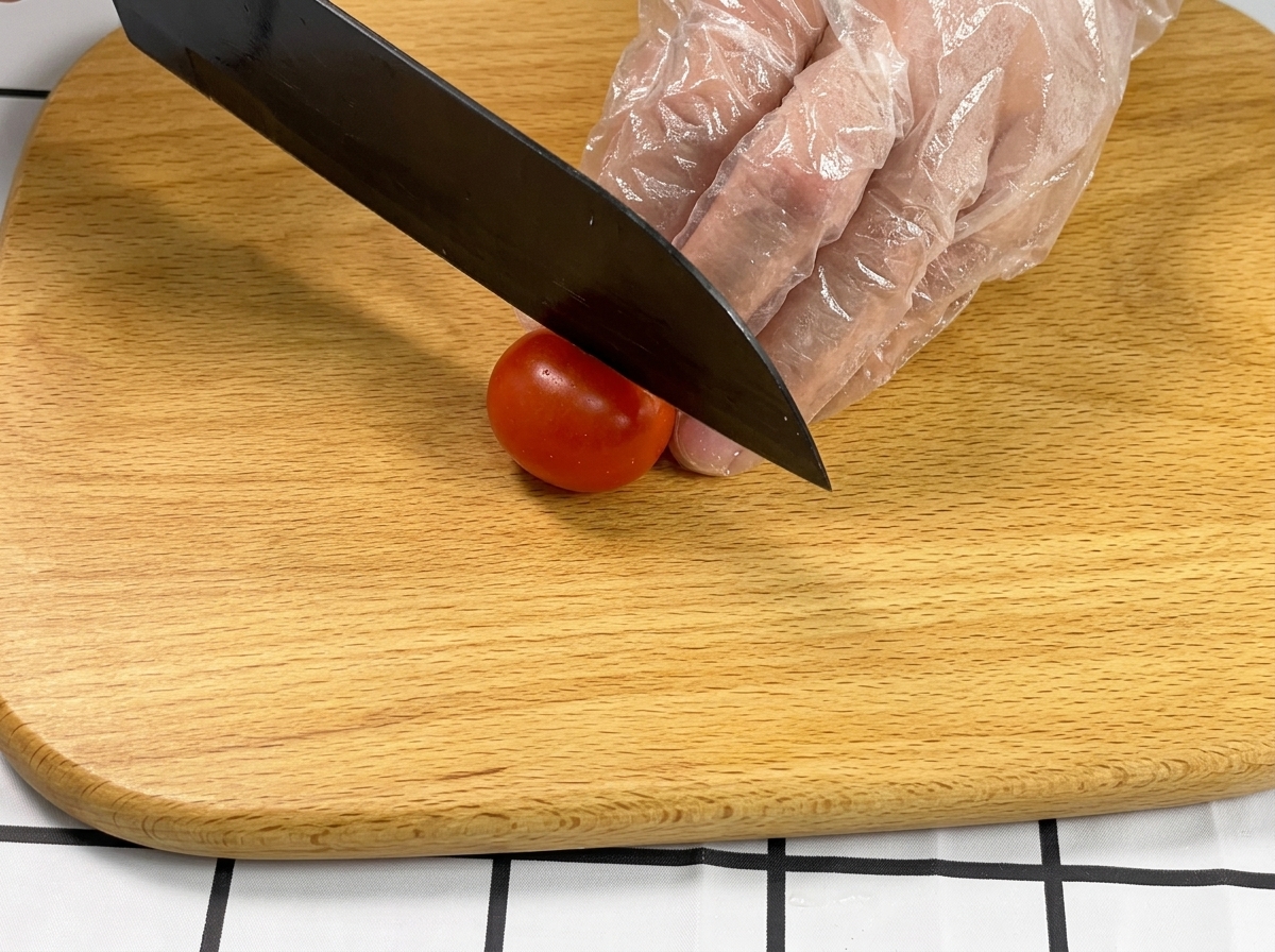 Eine Hand hält eine Kirschtomate fest, während ein schwarzes Messer sie auf einem Holzbrett halbiert.
