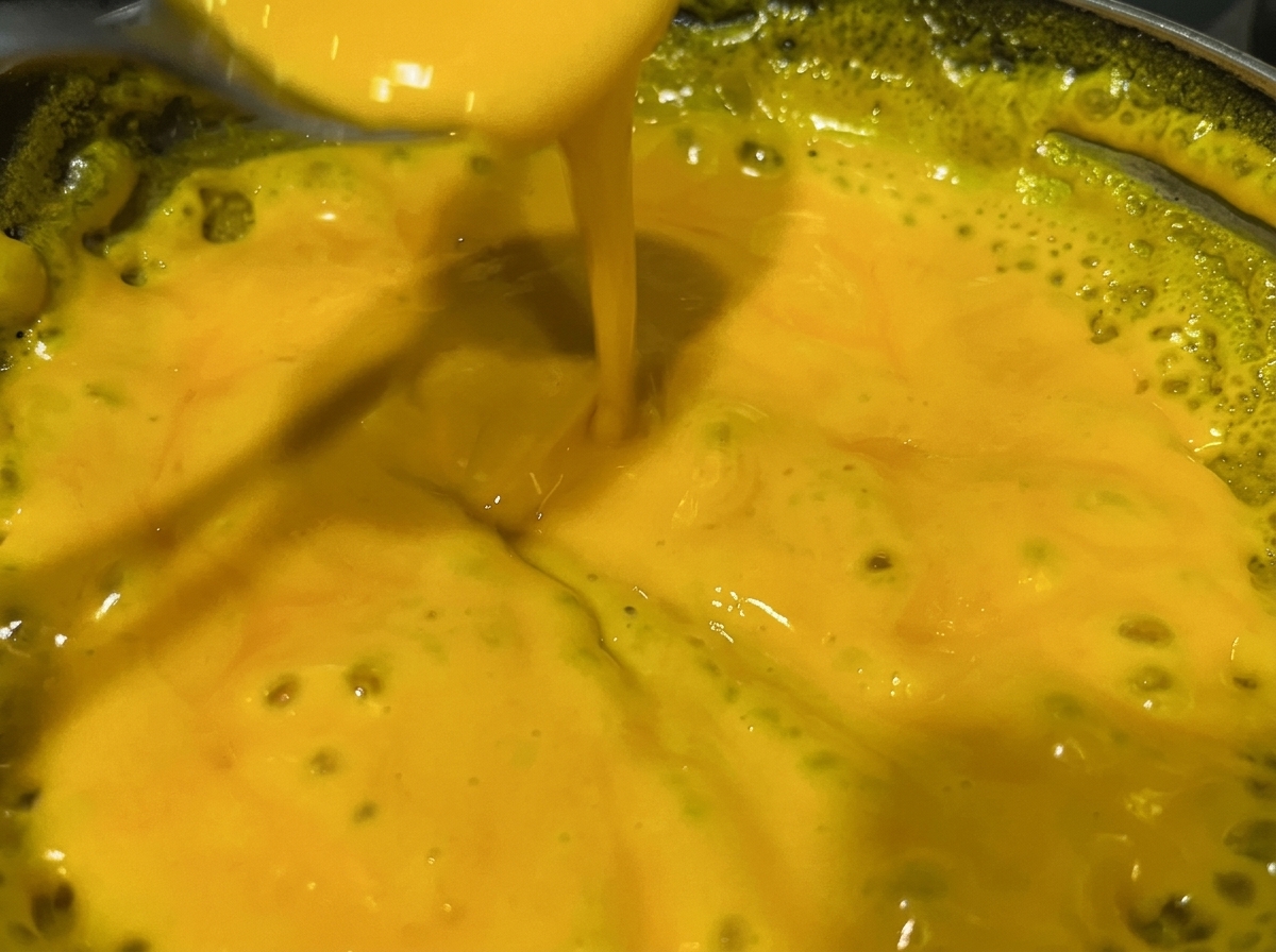Ein Löffel rührt in einer gelben, blubbernden Käsesauce in einem Metalltopf.