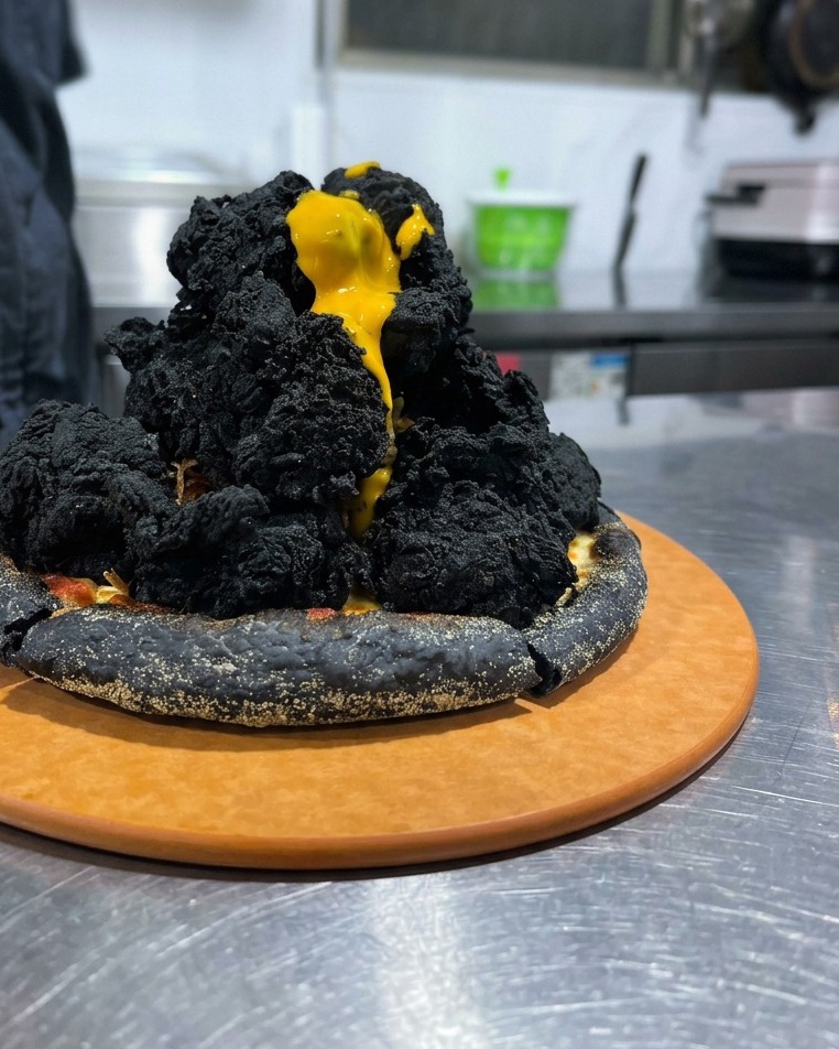Gelbe Käsesauce fließt wie Lava über den schwarzen Hähnchenberg auf der Pizza.