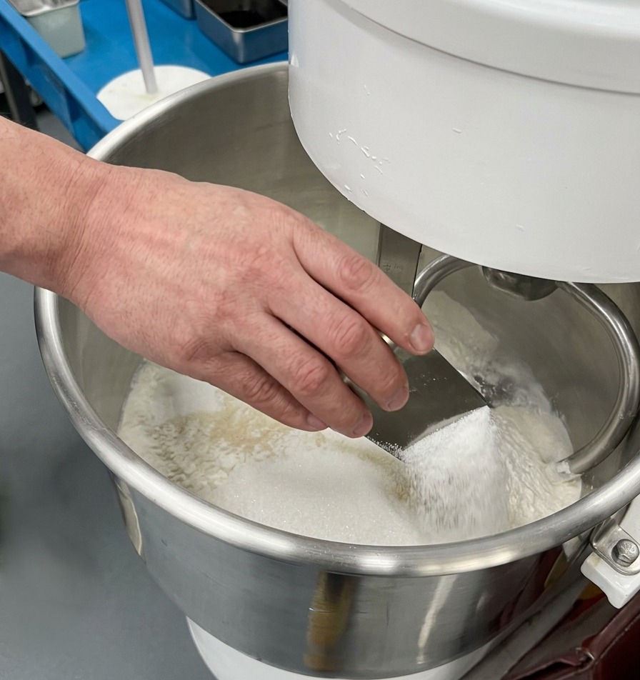 Eine Hand schüttet weißen Zucker aus einer kleinen Metallschaufel in eine Schüssel mit Mehl, Hefe und Salz.