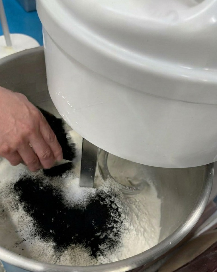 Eine Hand schüttet feines schwarzes Pulver in eine Rührschüssel mit Mehl und anderen trockenen Zutaten.