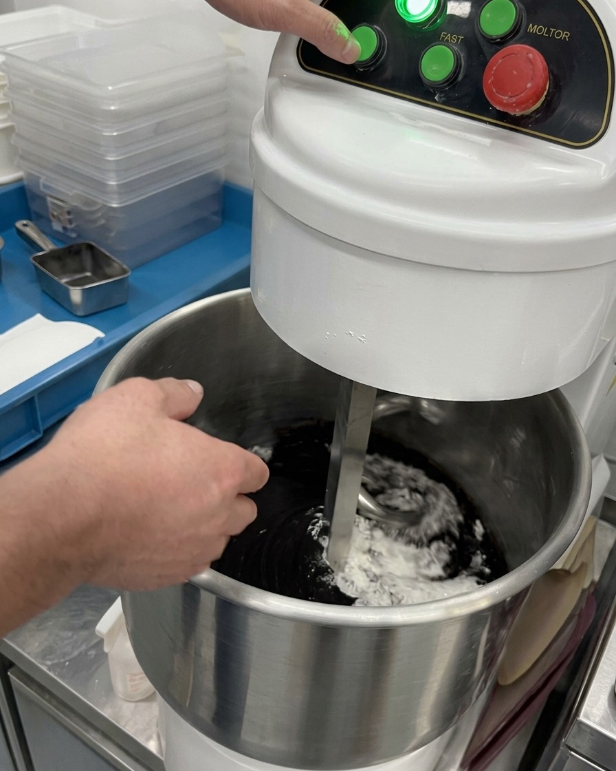 Eine Hand drückt den grünen Startknopf einer Knetmaschine, während der Knethaken schwarze und weiße trockene Zutaten vermischt.