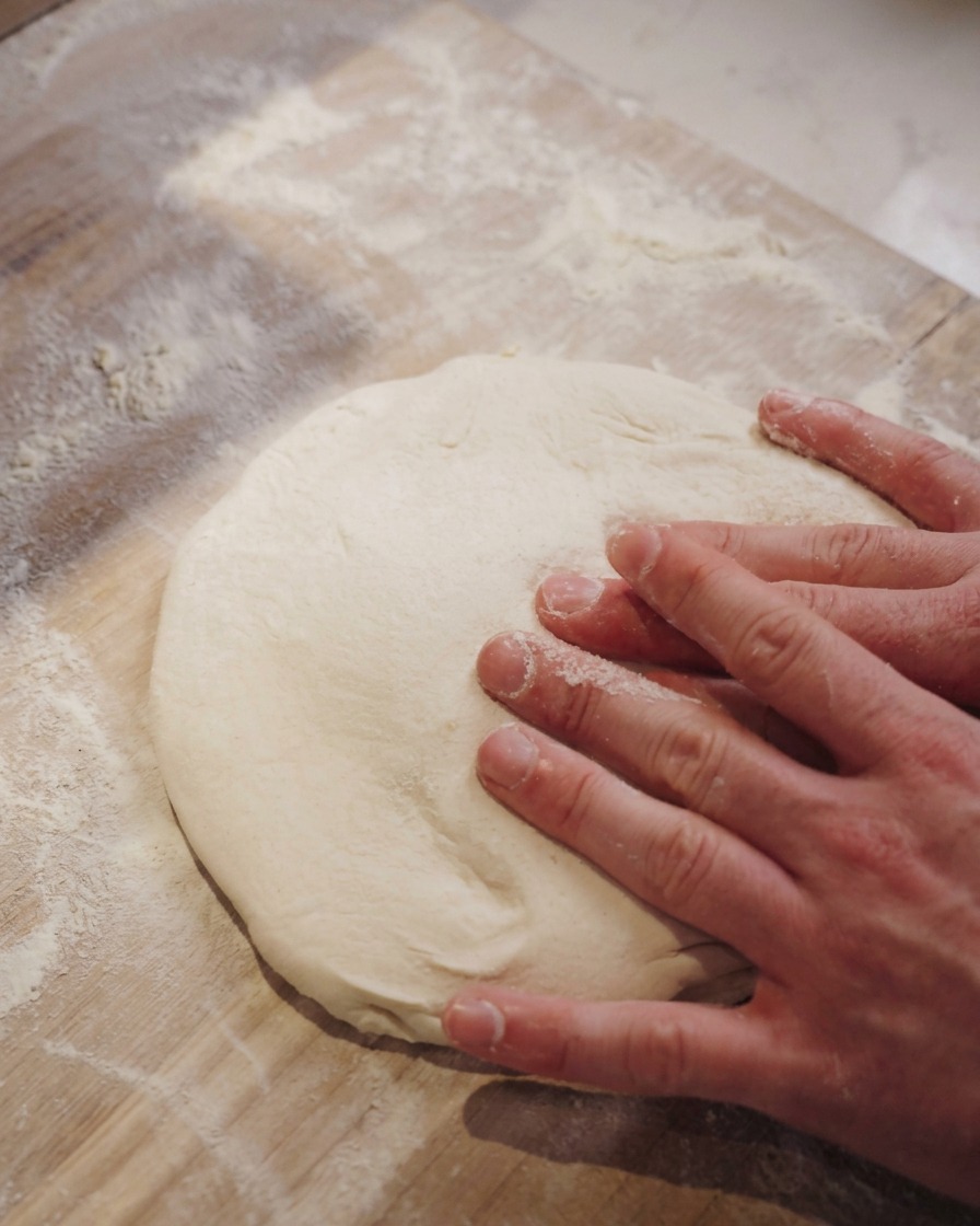 Zwei Hände drücken die Mitte eines weichen Pizzateigs auf einer bemehlten Holzfläche flach.