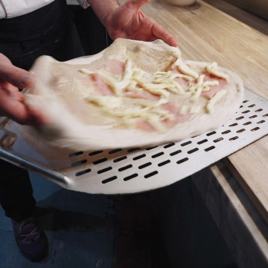 Ein Koch hält die vorbereitete Pizza auf einer Metallschaufel, belegt mit Creme, Schinken und Käsestreifen.