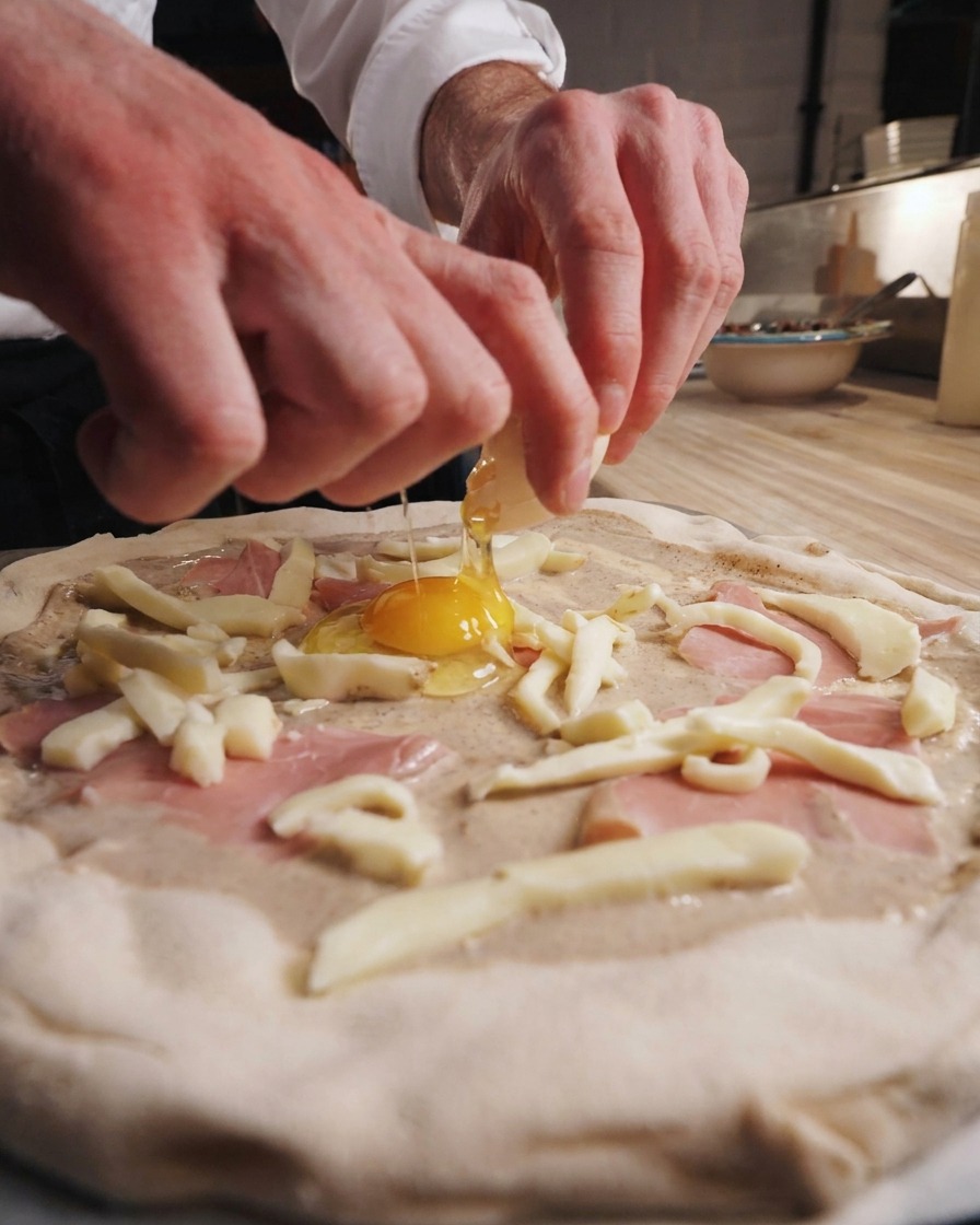 Ein Ei wird direkt in das Zentrum der reich belegten Pizza aufgeschlagen.