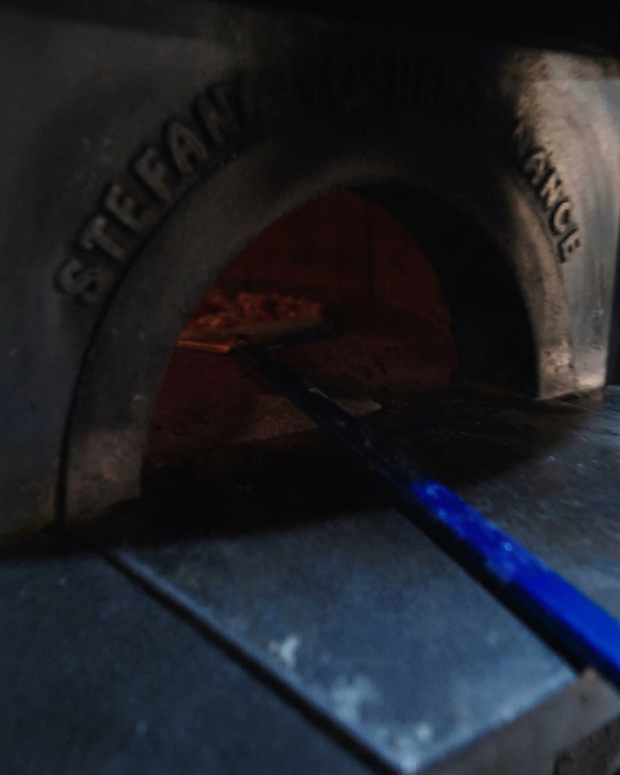 Eine blaue Pizzaschaufel schiebt die Pizza in den glühenden Holzofen.