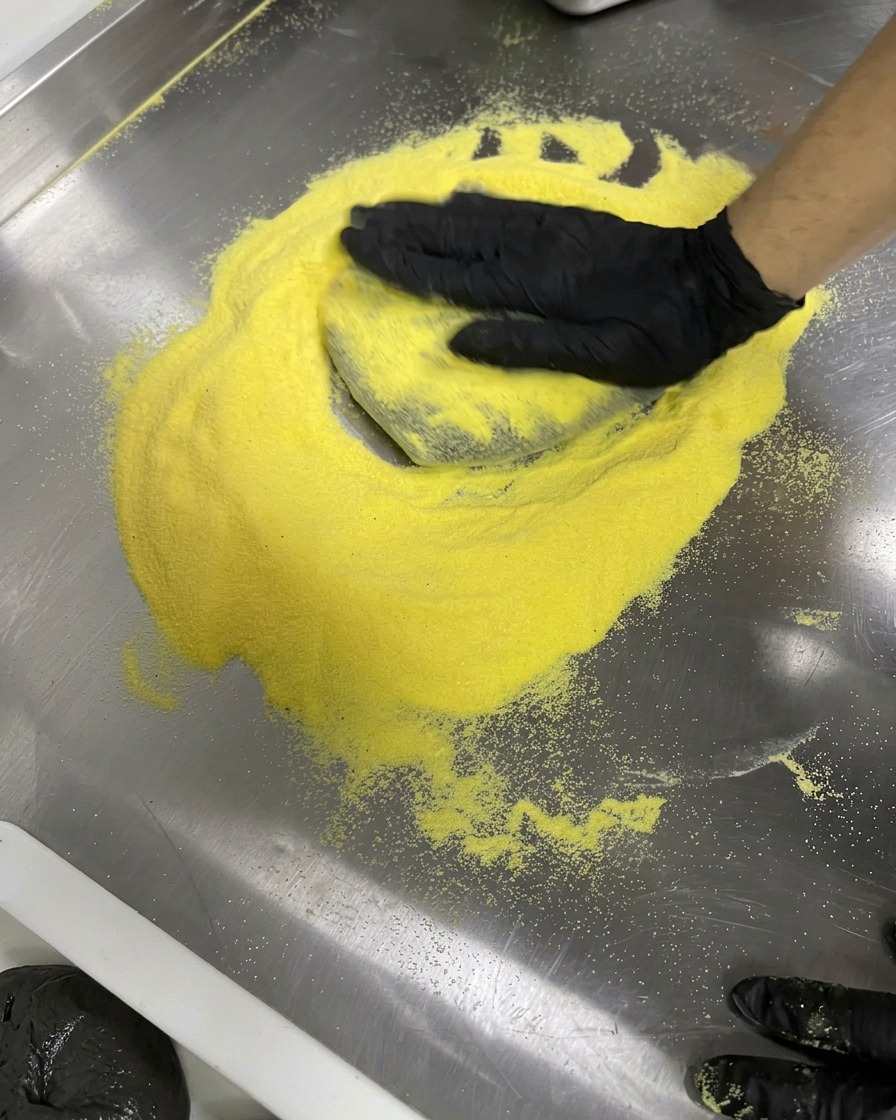 Hände in Handschuhen dehnen schwarzen Teig kreisförmig auf einer mit gelbem Mehl bestäubten Stahlfläche.