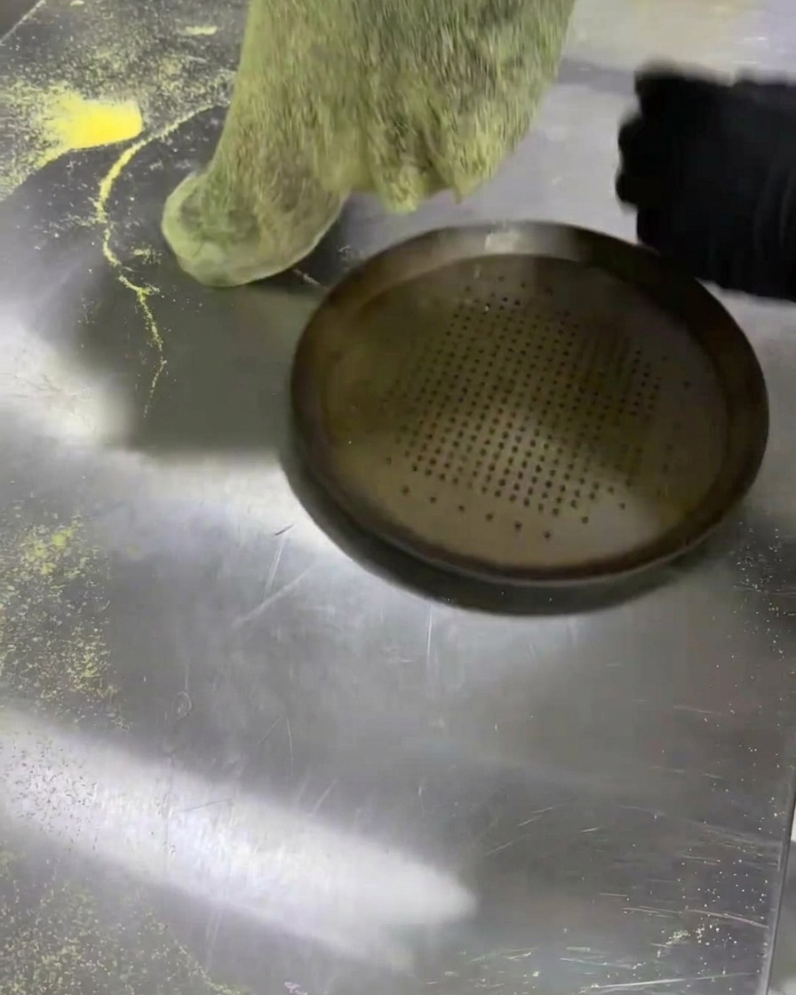 Ein flacher schwarzer Teigfladen wird auf ein perforiertes Pizzablech gelegt.