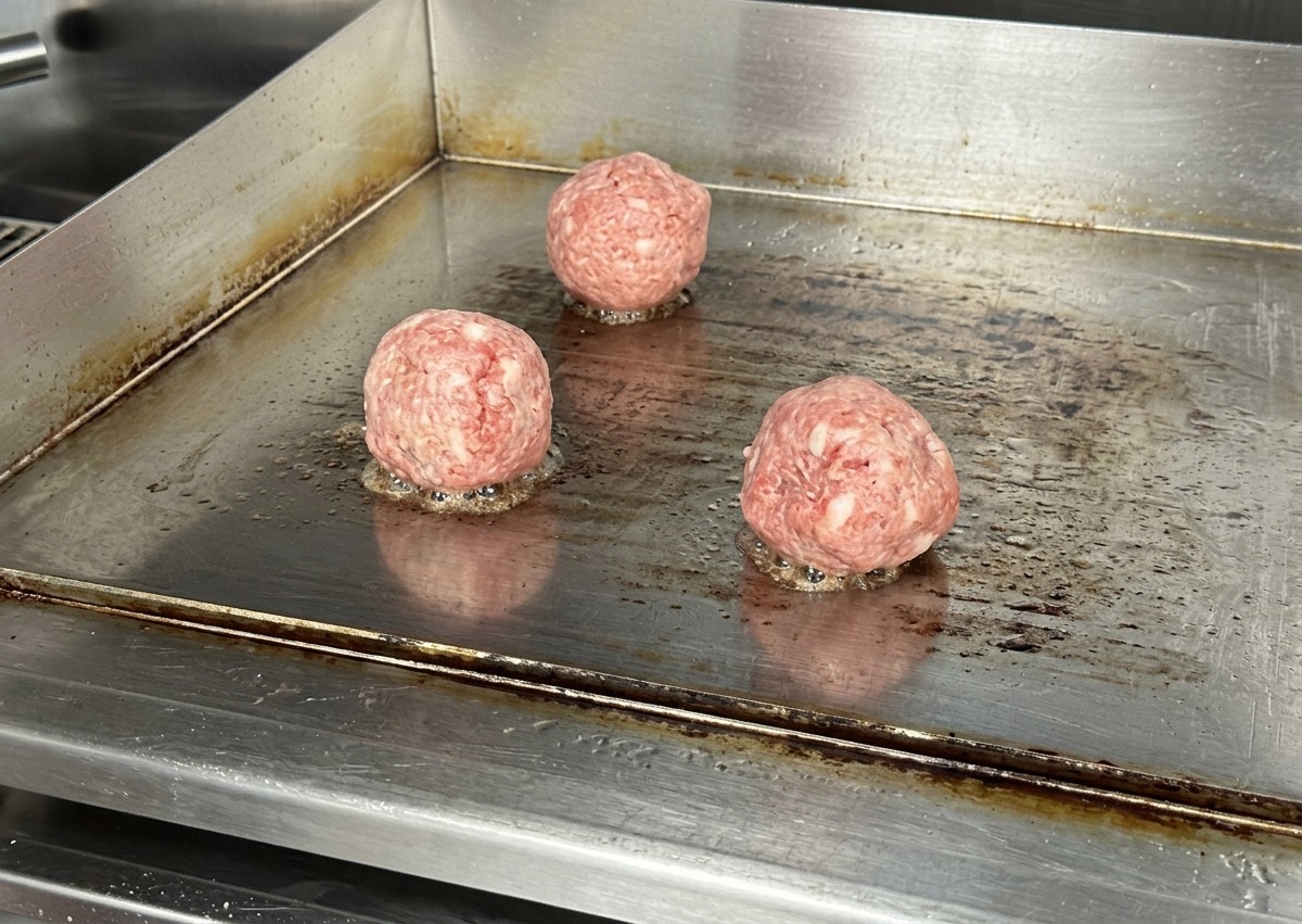 Drei runde, rohe Rinderhackkugeln liegen auf einer heißen Edelstahl-Grillplatte mit leichtem Zischen am Boden.