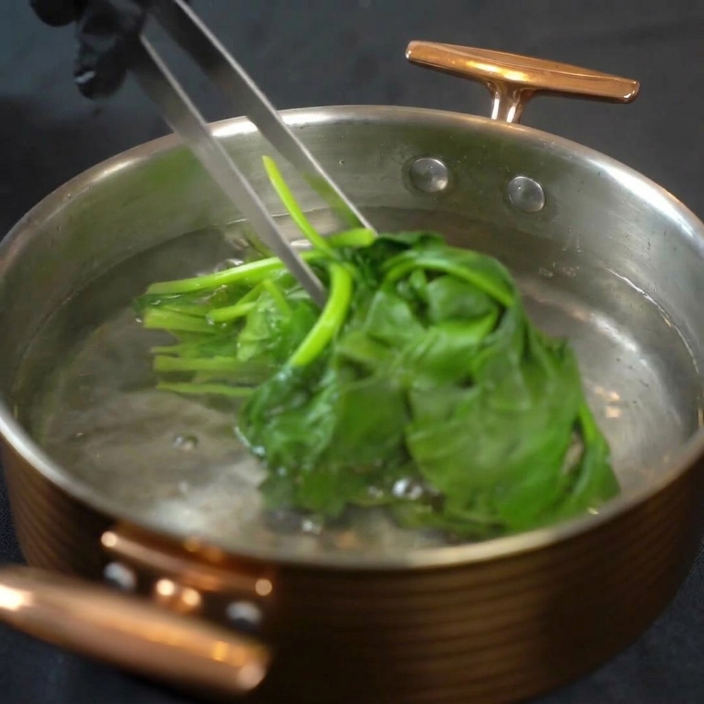 Eine Metallzange hebt hellgrüne blanchierte Spinatblätter aus einem Topf mit kochendem Wasser.