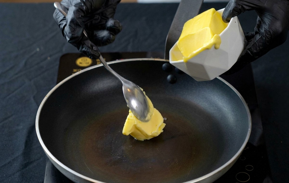 Ein Löffel gibt ein Stück Butter in eine erhitzte schwarze Bratpfanne.