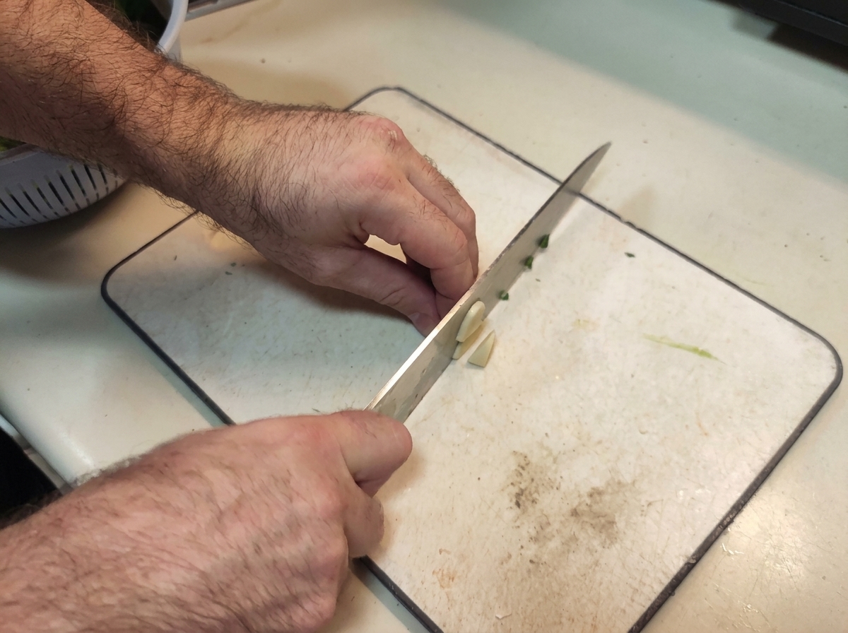 Hände, die mit einem Kochmesser Knoblauchzehen auf einem weißen Schneidebrett fein würfeln.