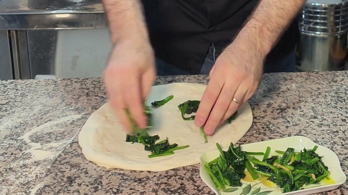 Hände verteilen den gegarten grünen Spinat gleichmäßig auf einem handgeformten Pizzateig.
