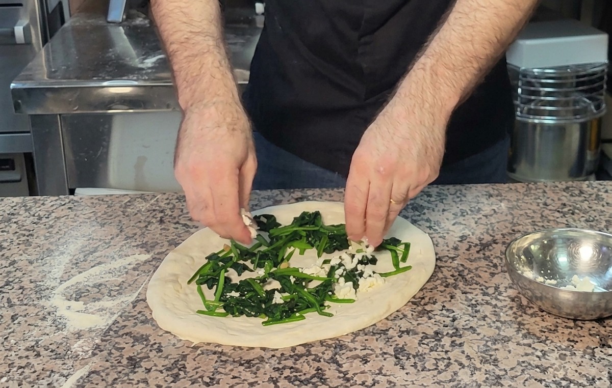Hände bröseln Feta-Käse über eine Schicht Spinat auf dem Pizzateig.
