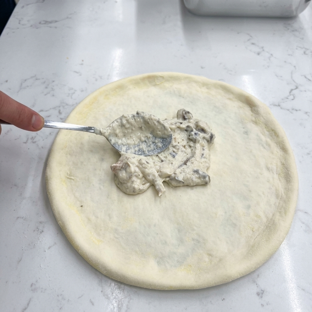 Eine Hand verstreicht mit einem Metalllöffel weiße, cremige Pilzsauce auf dem rohen Pizzateig.