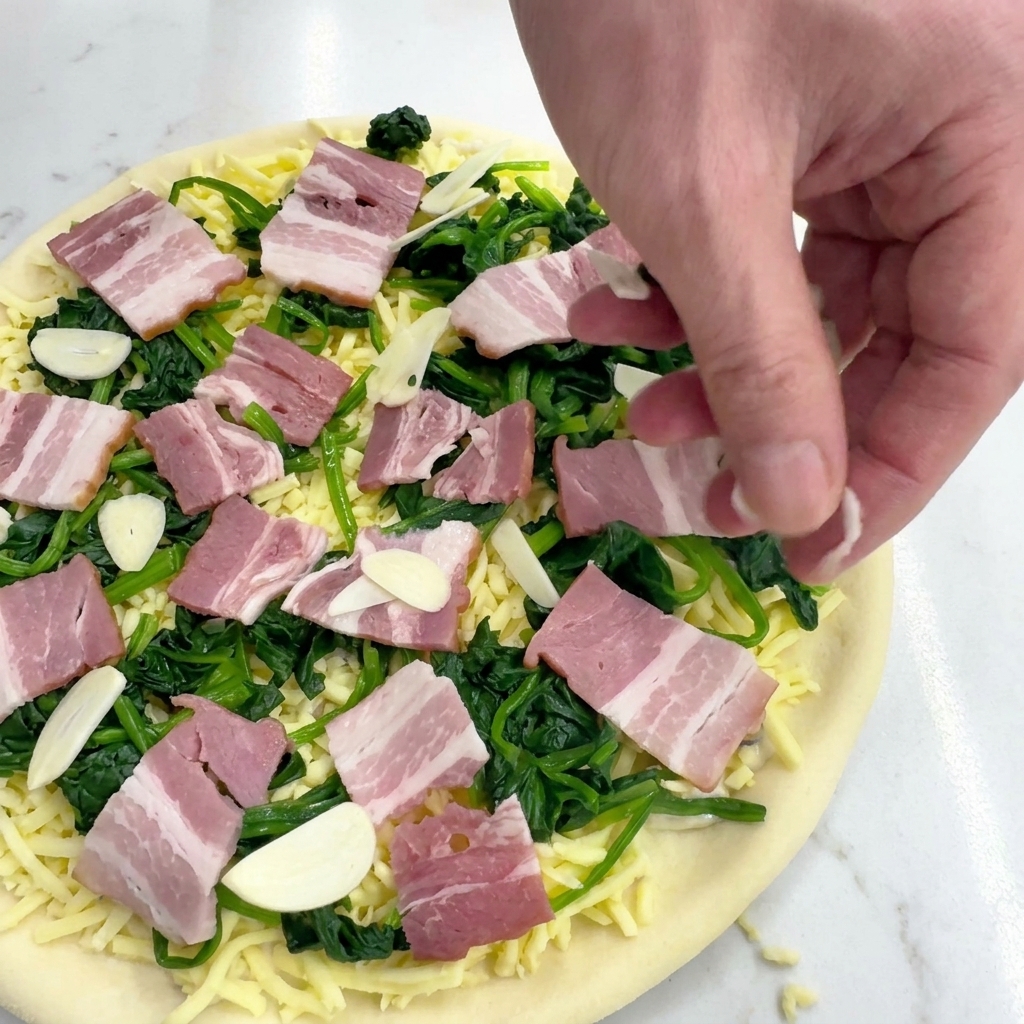 Eine Hand streut dünne Knoblauchscheiben über die Pizza mit Speck und Spinat.