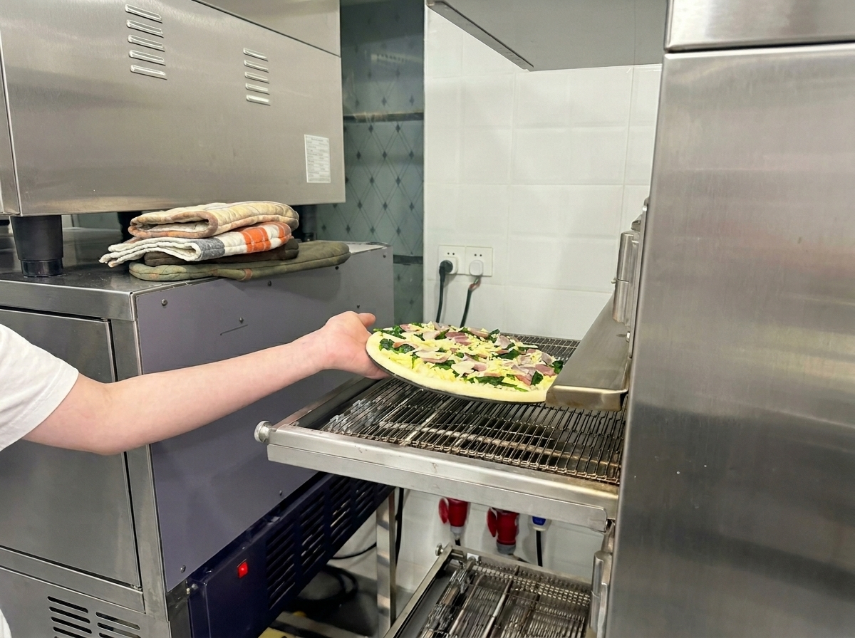 Ein Arm schiebt die vorbereitete Pizza auf das Laufband eines professionellen Pizzaofens.