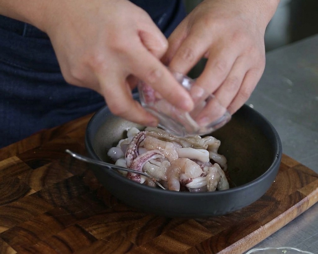 Hände geben rohe Tintenfischringe und Garnelen in eine dunkle Rührschüssel mit Sauce und anderen Meeresfrüchten.