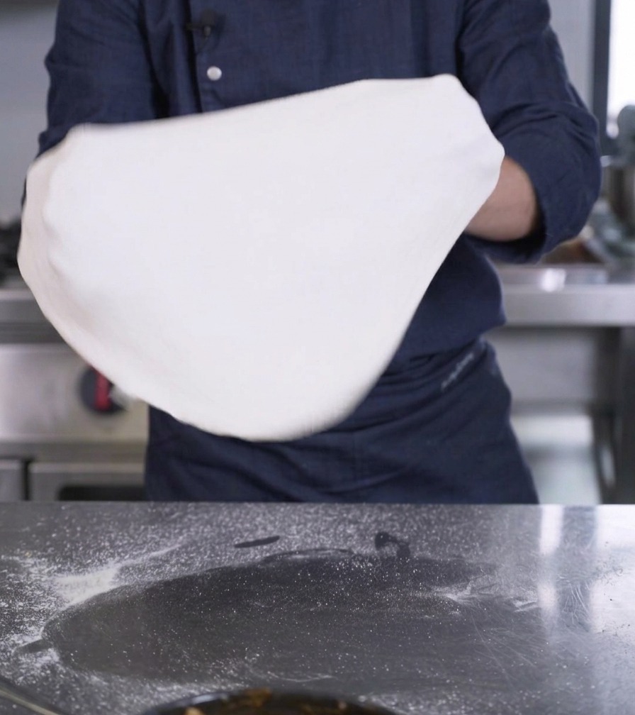 Die Hände einer Person halten und dehnen eine große, dünne Scheibe weißen Pizzateigs in der Luft.