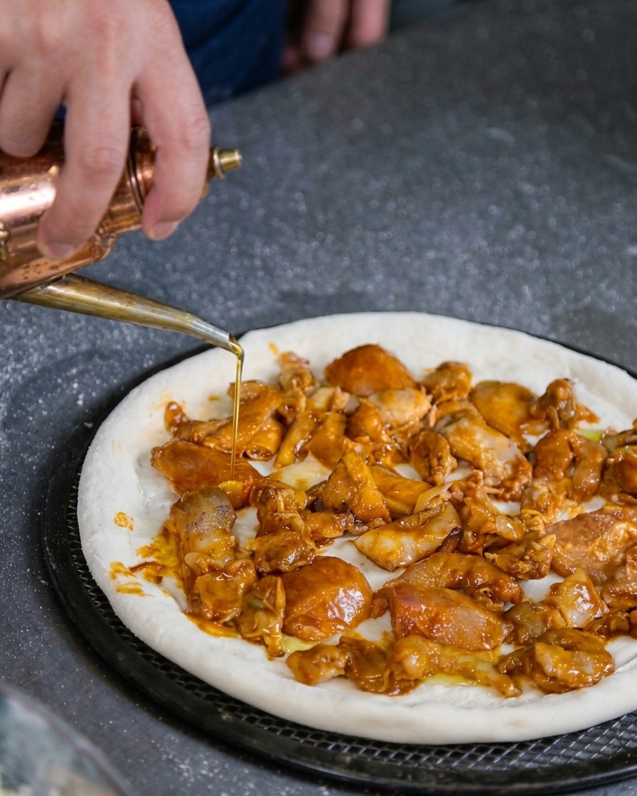 Eine Hand beträufelt mit einem Kupfer-Ölspender vorsichtig eine ungebackene Pizza, die mit marinierten Meeresfrüchten belegt ist.