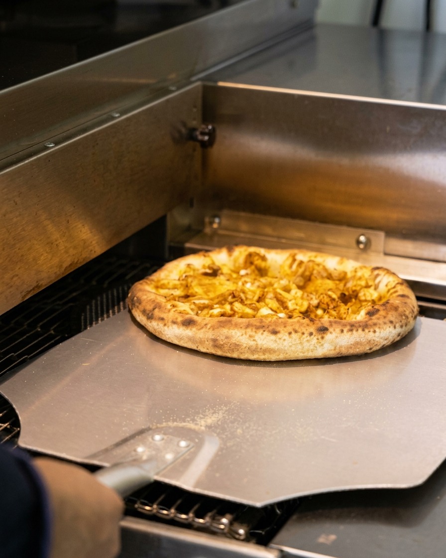 Eine frisch gebackene Meeresfrüchte-Pizza mit dickem, leicht verkohltem Rand wird mit einem Metall-Pizzaschieber aus einem gewerblichen Ofen geholt.