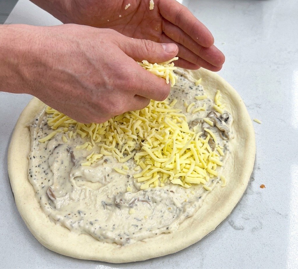 Zwei Hände streuen großzügig geriebenen Mozzarella über den mit Sauce bestrichenen Pizzaboden.