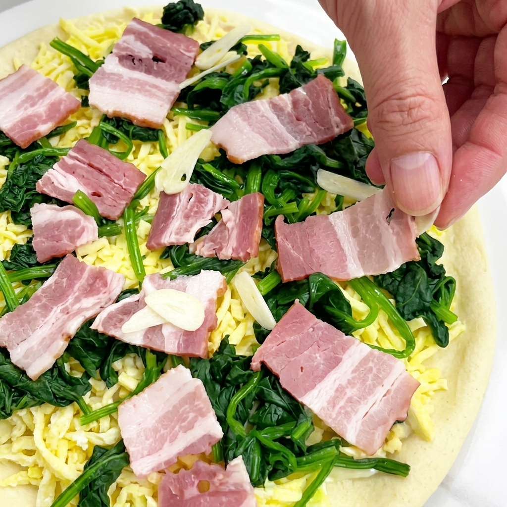 Eine Hand streut dünne Knoblauchscheiben über die vorbereitete Pizza mit Speck und Spinat.