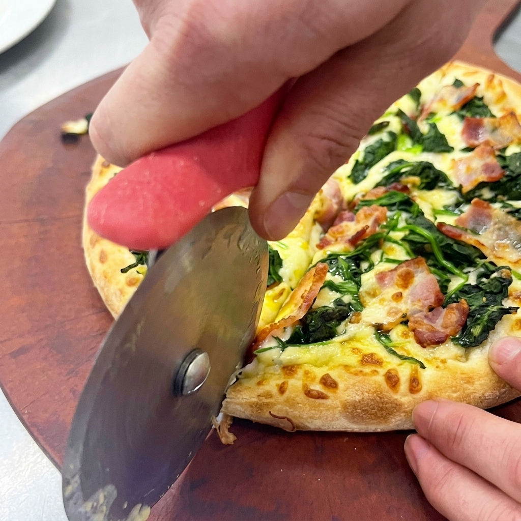 Hände schneiden eine frisch gebackene Spinat-Speck-Pizza mit einem Rollschneider auf einem Holzbrett.