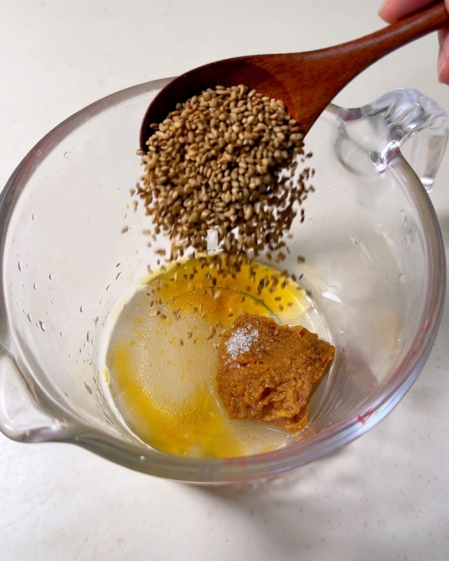 Ein Holzlöffel gibt gerösteten Sesam in einen Messbecher mit Misopaste, Zucker und Sesamöl.