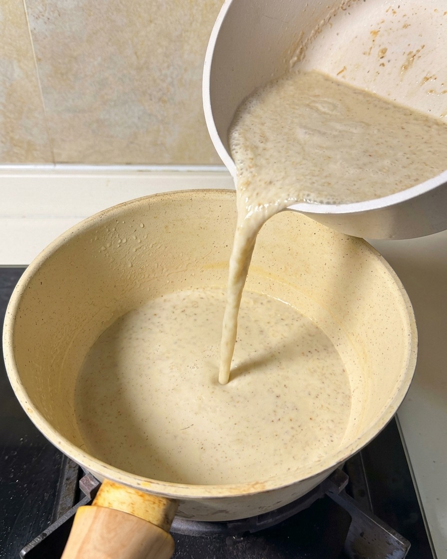 Eine beige Suppenmischung wird aus einem Krug in einen kleinen Topf auf dem Herd gegossen.