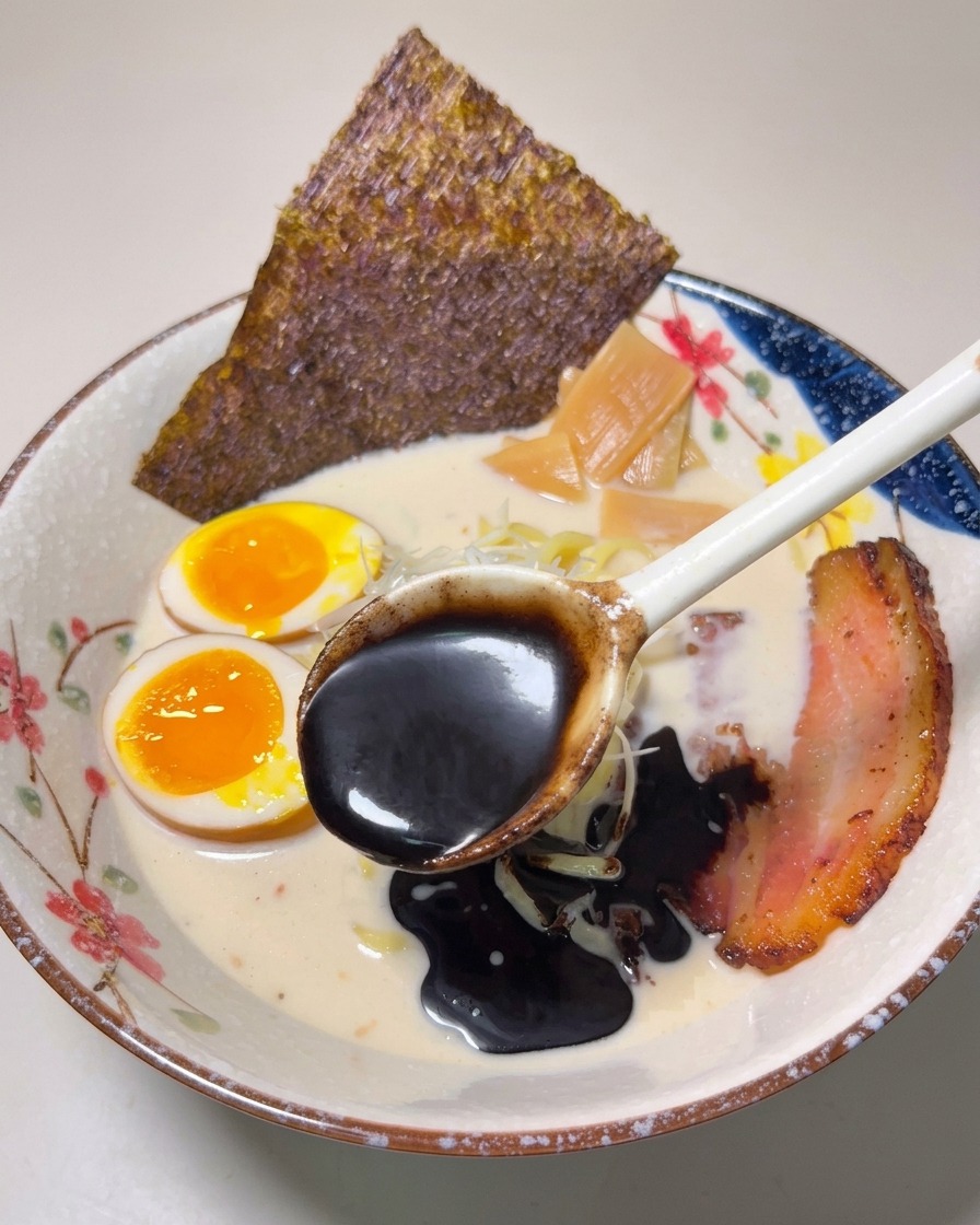 Ein Löffel mit tiefschwarzem Knoblauchöl wird über die fertige Ramen-Schüssel geträufelt.