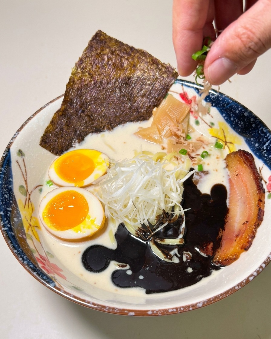 Eine Hand streut gehackte Frühlingszwiebeln über die fertig angerichtete Schüssel Ramen.