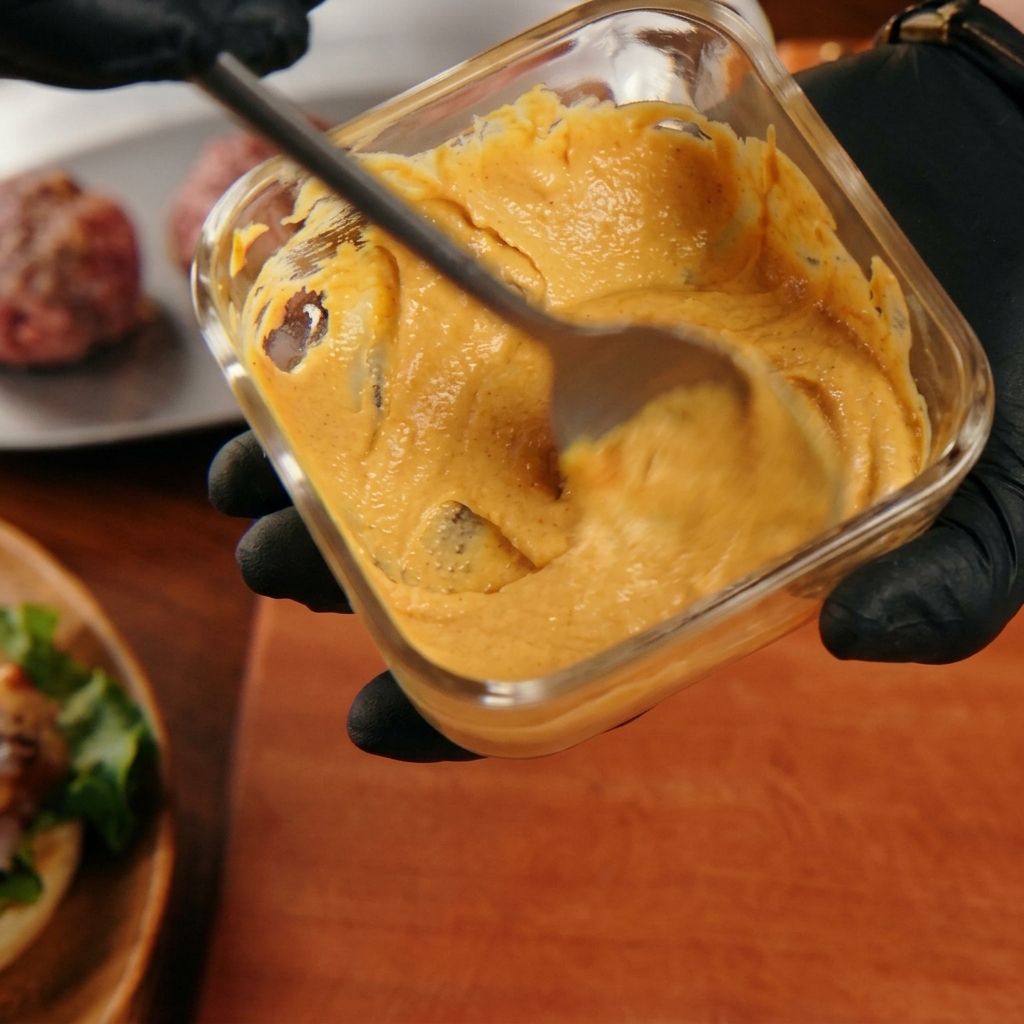 Eine Hand rührt eine cremige, orangefarbene Burgersauce mit einem Löffel in einem Glasbehälter an.