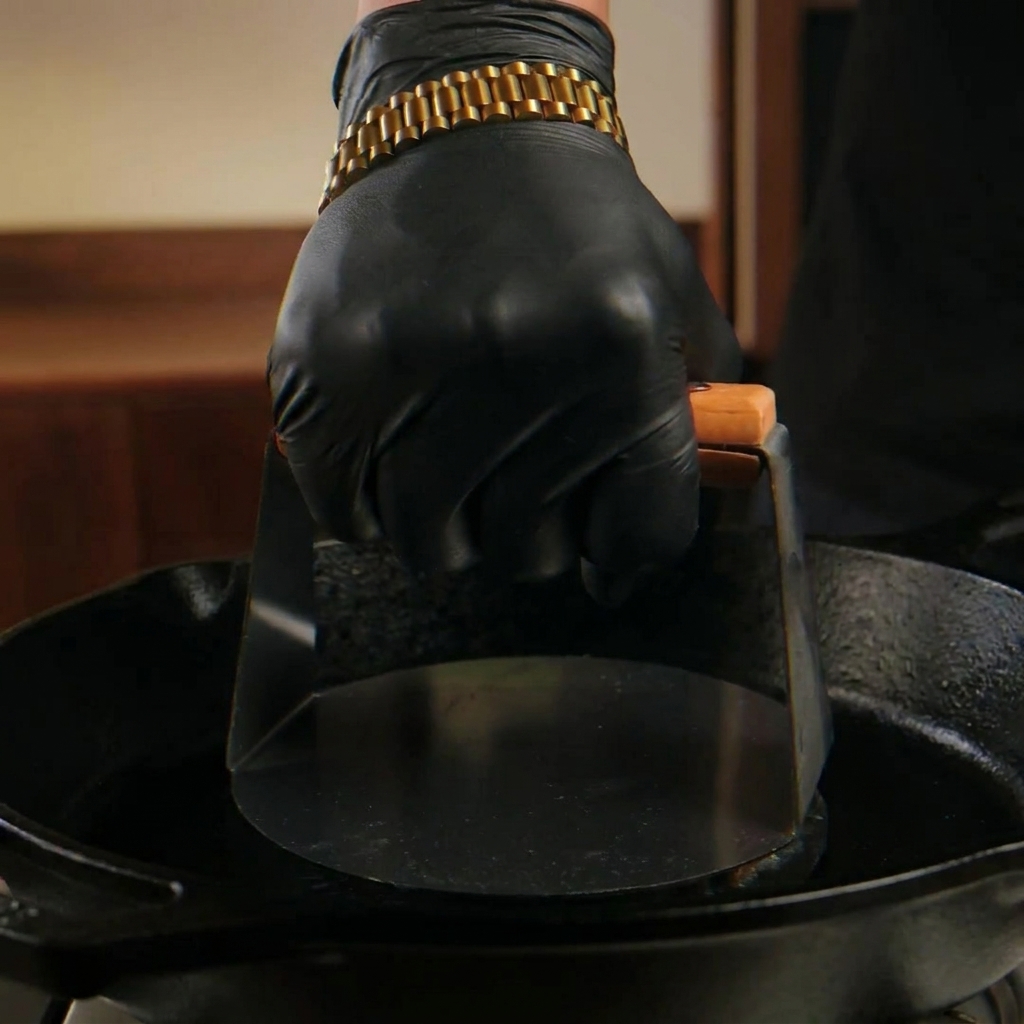 Eine Hand mit schwarzem Handschuh drückt eine Fleischkugel mit einer schweren Metallpresse in einer rauchenden Gusseisenpfanne flach.