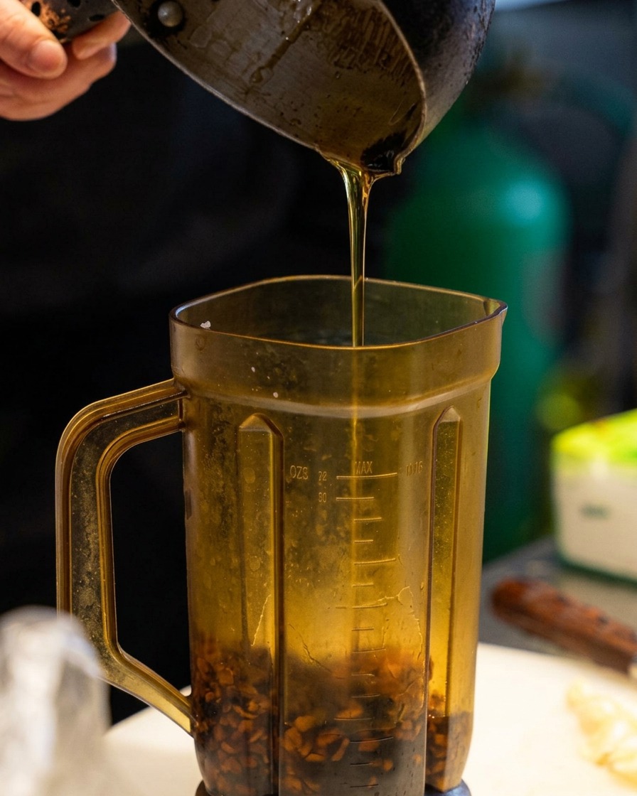 Goldenes Sesamöl wird über die dunklen Knoblauchstücke in den Mixbecher gegossen.