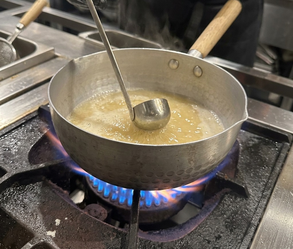 Dunkle Sojasauce wird aus einer kleinen Metallschüssel in die kochende Brühe gegeben.