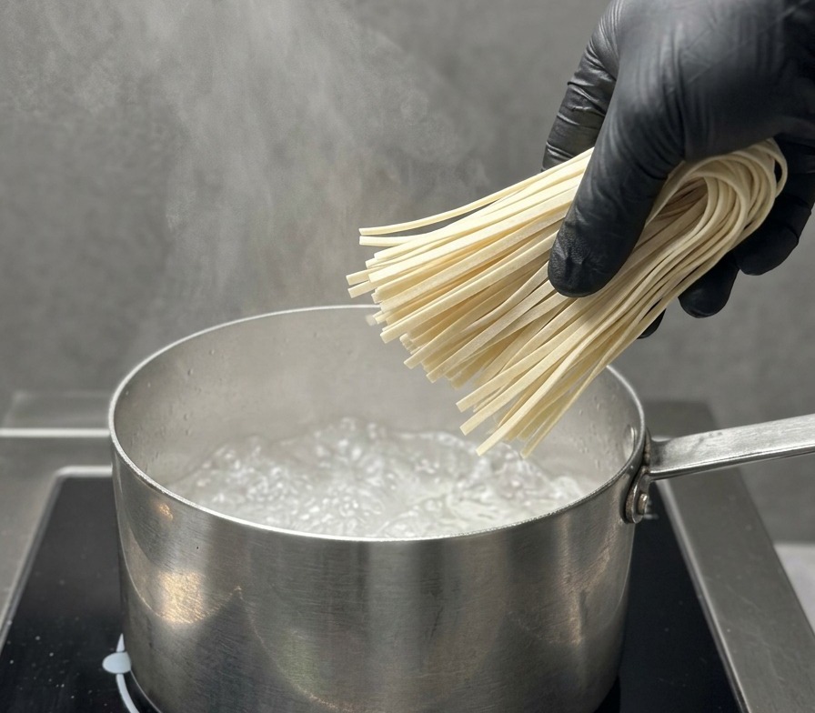 Eine Hand mit schwarzem Handschuh lässt ein Bündel trockene Ramen-Nudeln vorsichtig in kochendes Wasser fallen.