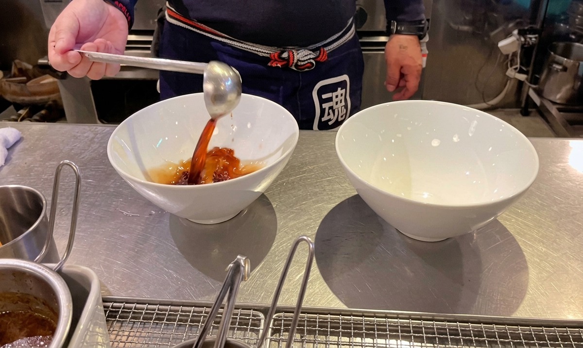 Ein Koch gießt dunkles Sojasaucen-Tare mit einer Schöpfkelle in weiße Ramen-Schüsseln.