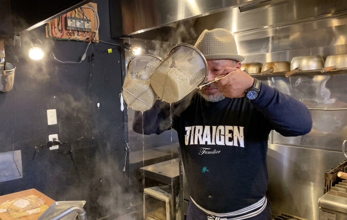 Ein Koch schüttelt Nudelsiebe kräftig über Kopfhöhe, um das Wasser von den gekochten Ramen zu entfernen.